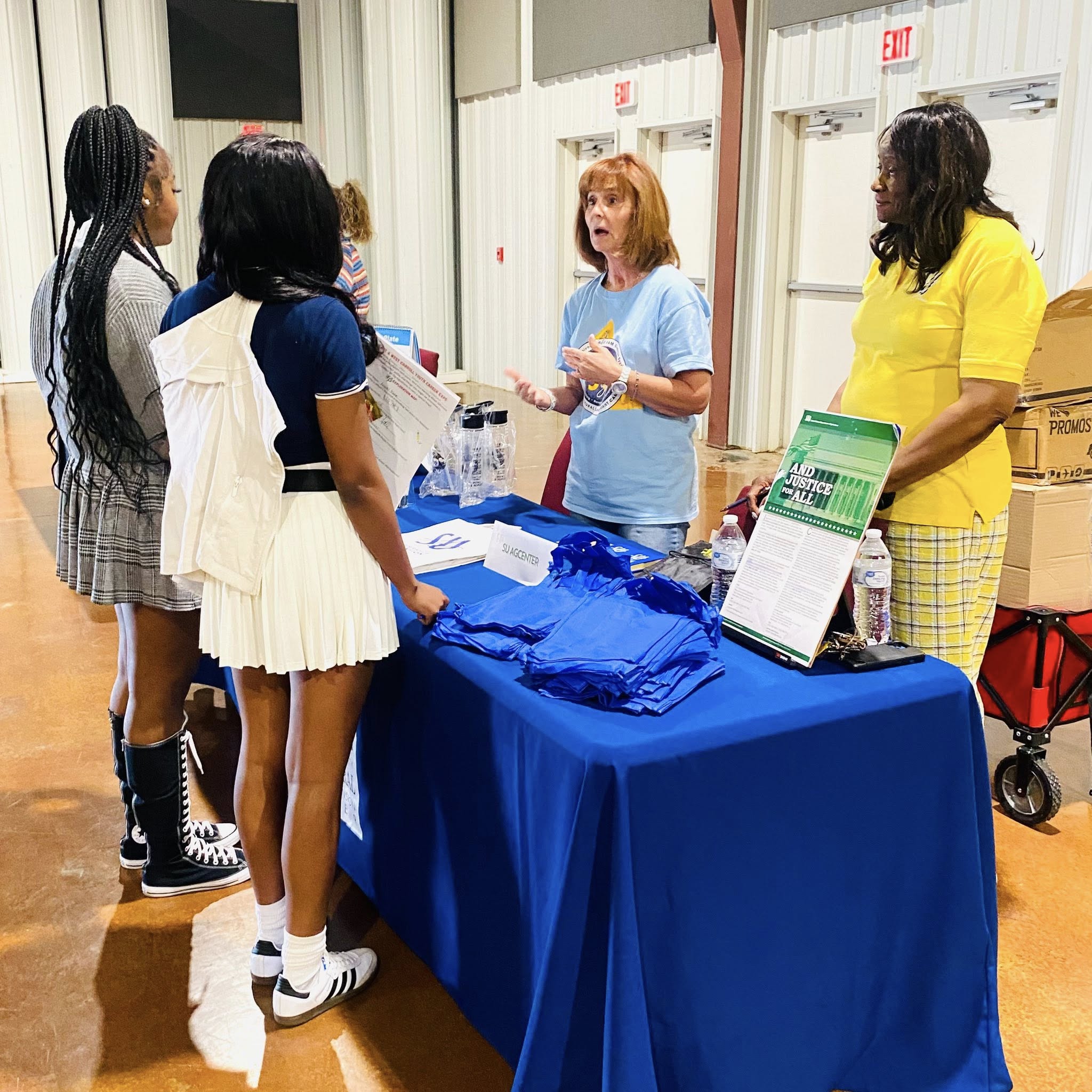 Two women behind a table talking to some teen girls.